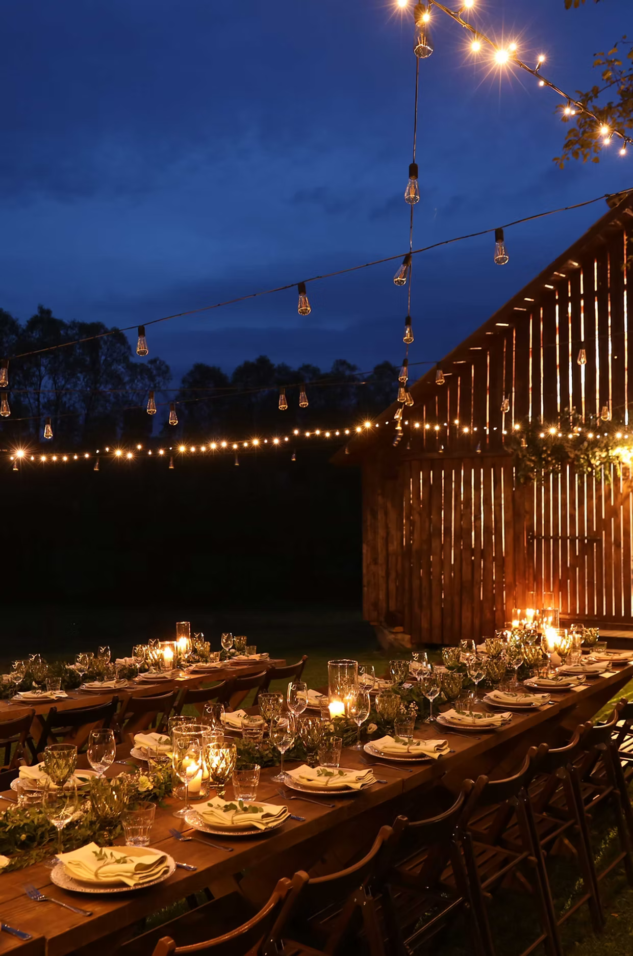 A long candle-lit dinner table set under string lights at dusk